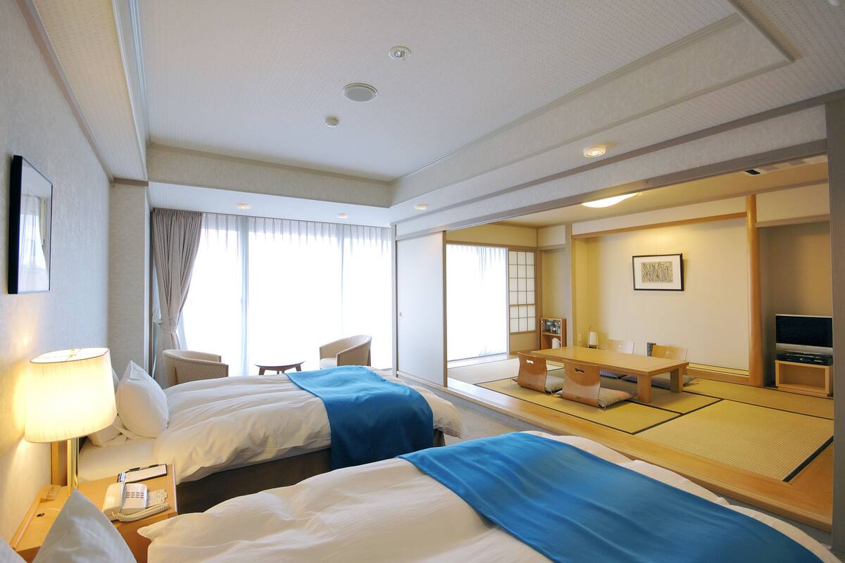 Une chambre d’hôtel avec deux lits, un coin salon avec une table et des chaises, un téléviseur et une grande fenêtre avec des rideaux.