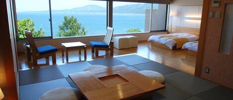 A modern hotel room with a large window offering a view of the sea and mountains. There is a bed, a seating area with a table, and a small sofa.