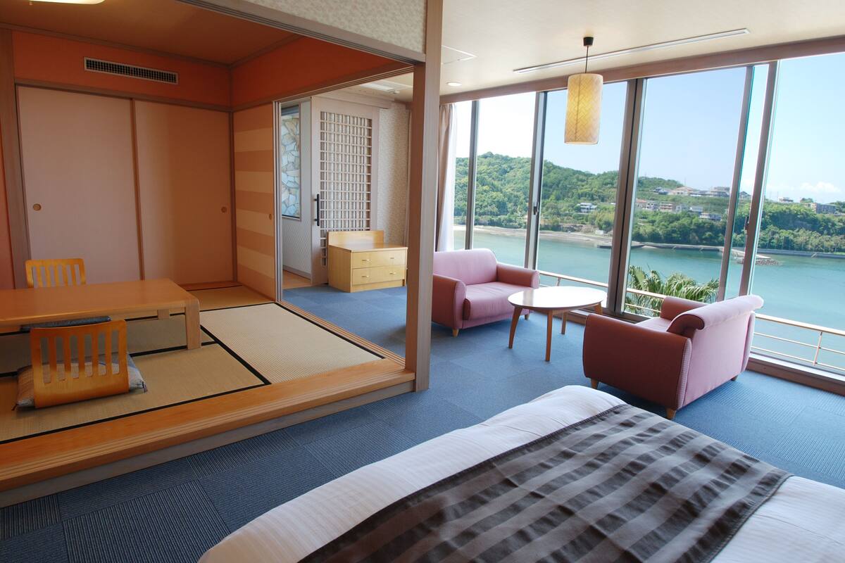 Une chambre d’hôtel comprenant un lit, une table à manger, une chaise, une petite table et un canapé. La fenêtre offre une vue sur une étendue d’eau et sur la verdure environnante.