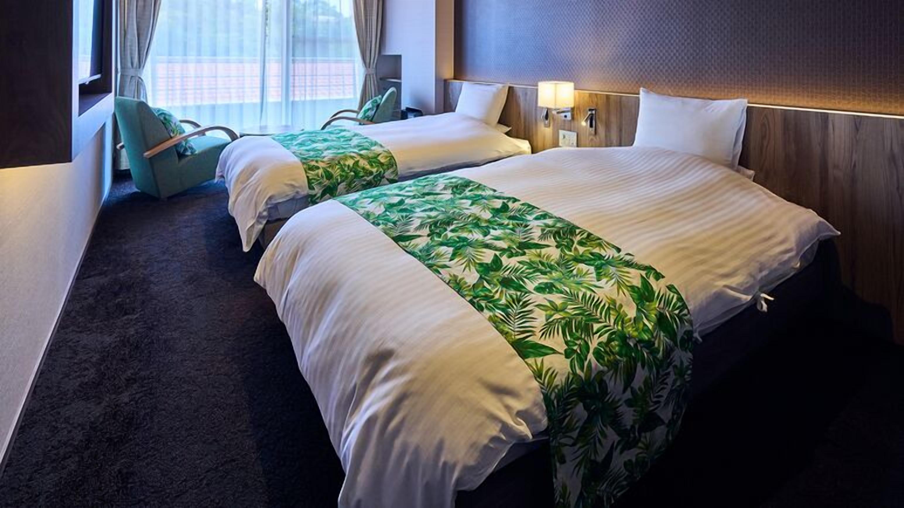 A hotel room with two beds, a window with curtains, a chair, and a wall-mounted lamp.