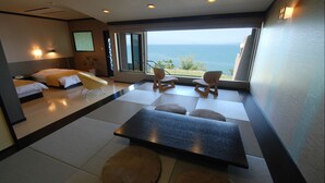 Quarto de hotel moderno com uma grande janela oferecendo vista para o mar, uma cama, área de estar com cadeiras e uma TV de tela plana.