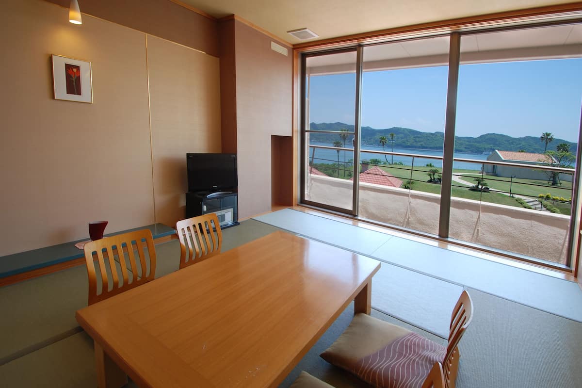 Une pièce comprenant une table à manger en bois, des chaises, un téléviseur et une grande fenêtre donnant sur la mer et les collines.