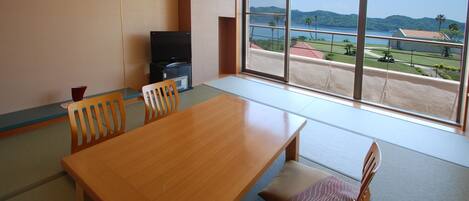 A room with a wooden dining table, chairs, a TV, and a large window offering a view of the sea and hills.