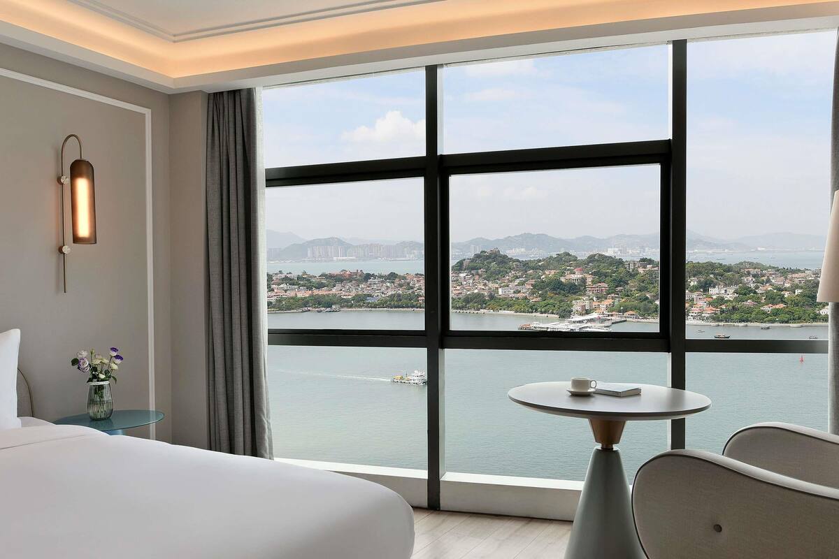 Une chambre d’hôtel avec une grande fenêtre, un lit, une table avec une tasse et une chaise.