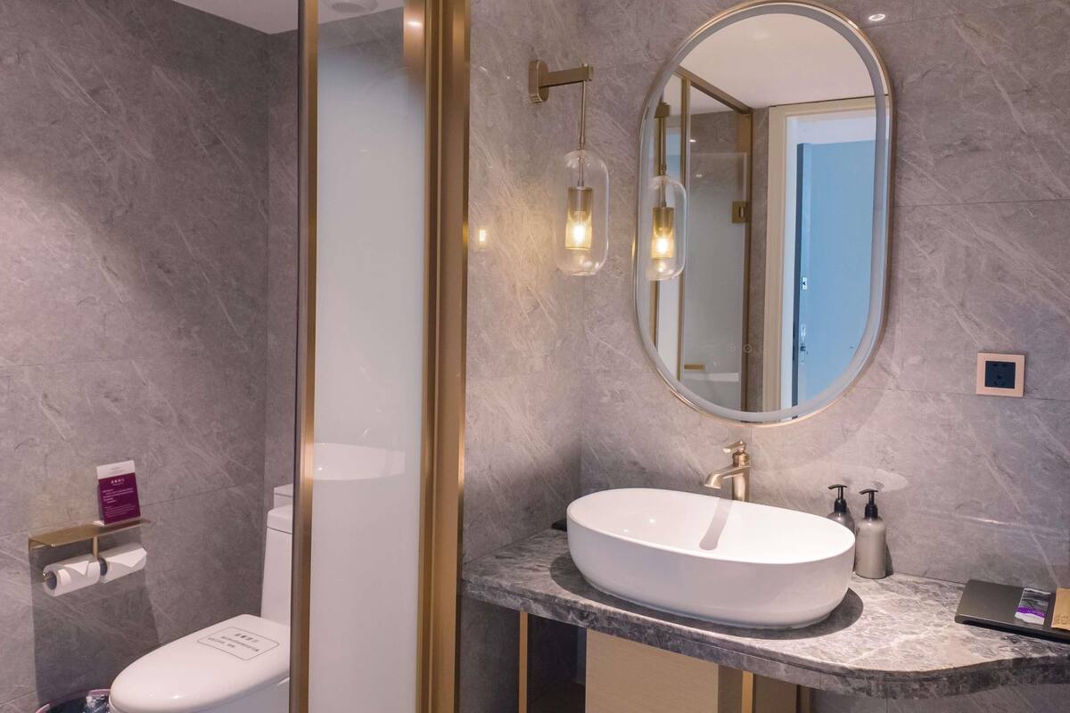 Une salle de bain moderne avec un mur en marbre, un grand lavabo ovale, un miroir encadré d’or et des toilettes dotées d’une poignée de chasse d’eau.