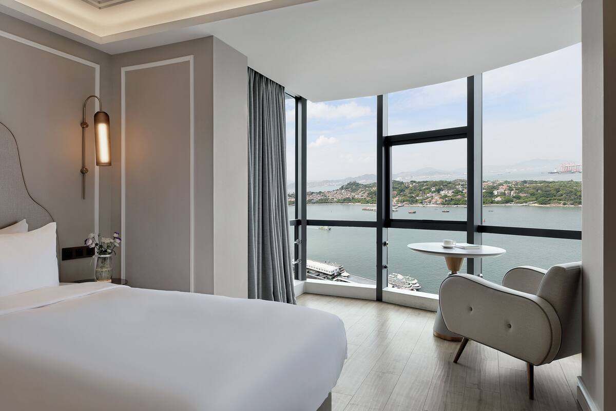 Une chambre d’hôtel avec un grand lit, une petite table, une chaise et une vue sur un plan d’eau et le paysage urbain.