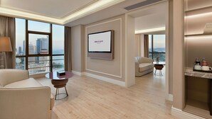 Habitación de hotel con un ventanal grande, un televisor de pantalla plana, un sofá, una mesa de centro y una mesita auxiliar.