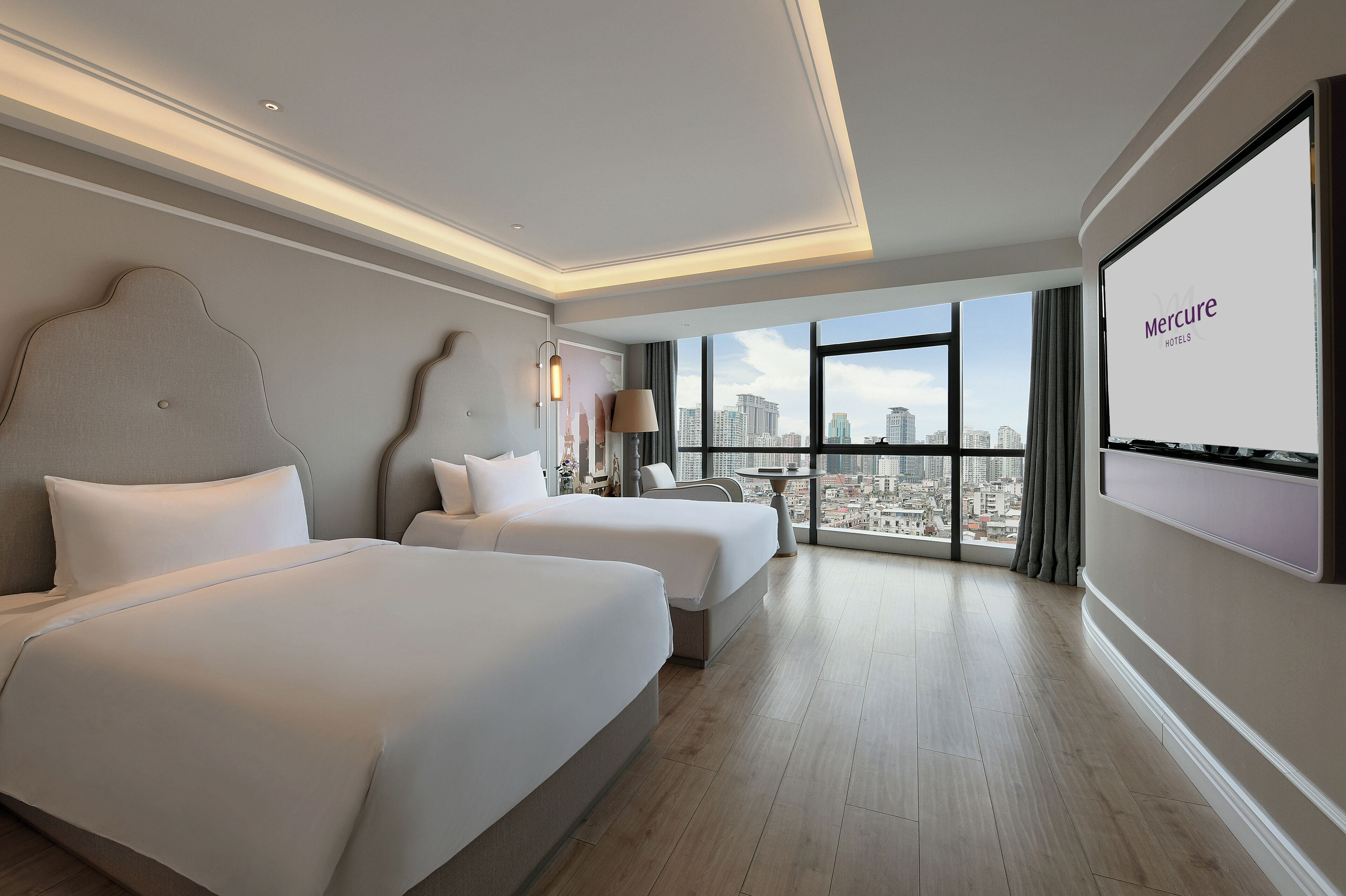 Habitación de hotel con dos camas, vistas a la ciudad y una pantalla mostrando 'Mercure Hotels'.