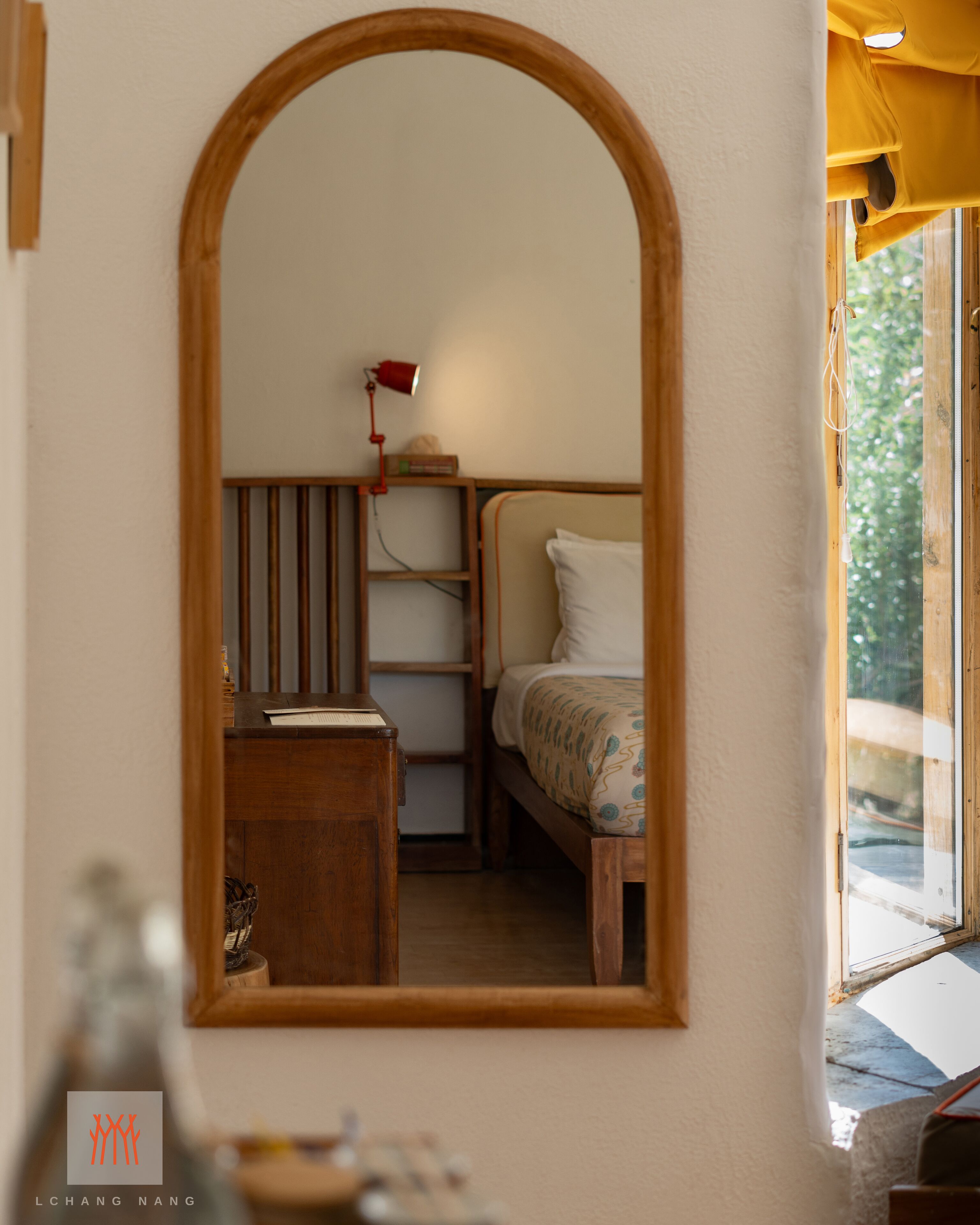 A room with a wooden-framed mirror, a bed with a patterned bedspread, a bedside table with a lamp, and a window with a view of greenery.