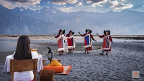 A group of people in traditional attire performing on a beach with a mountainous backdrop.