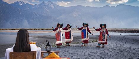 A group of people in traditional attire performing on a beach with a mountainous backdrop.
