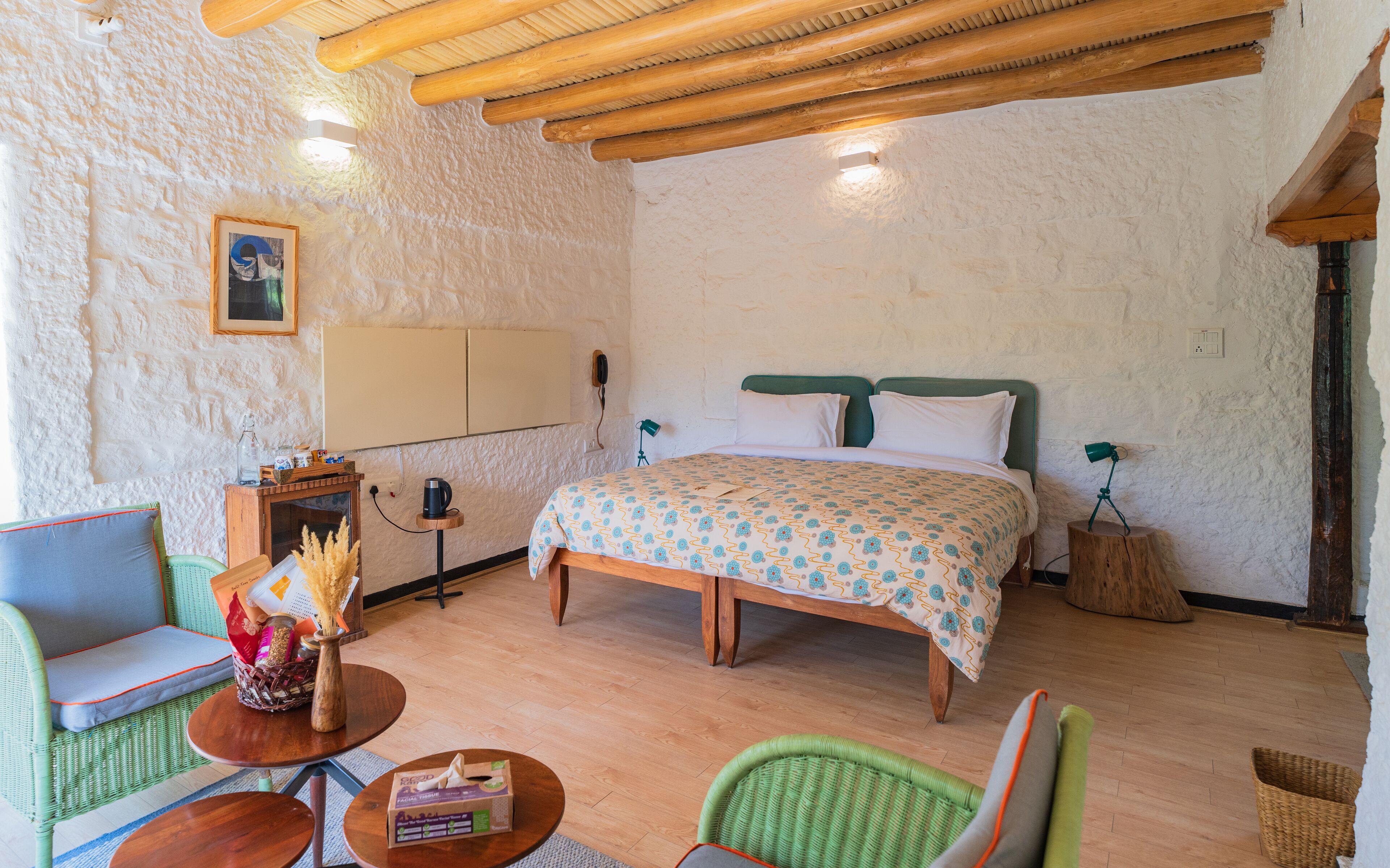 A cozy room with a bed, two green wicker chairs, a small wooden table, and a wall-mounted shelf with a vase and a bottle.