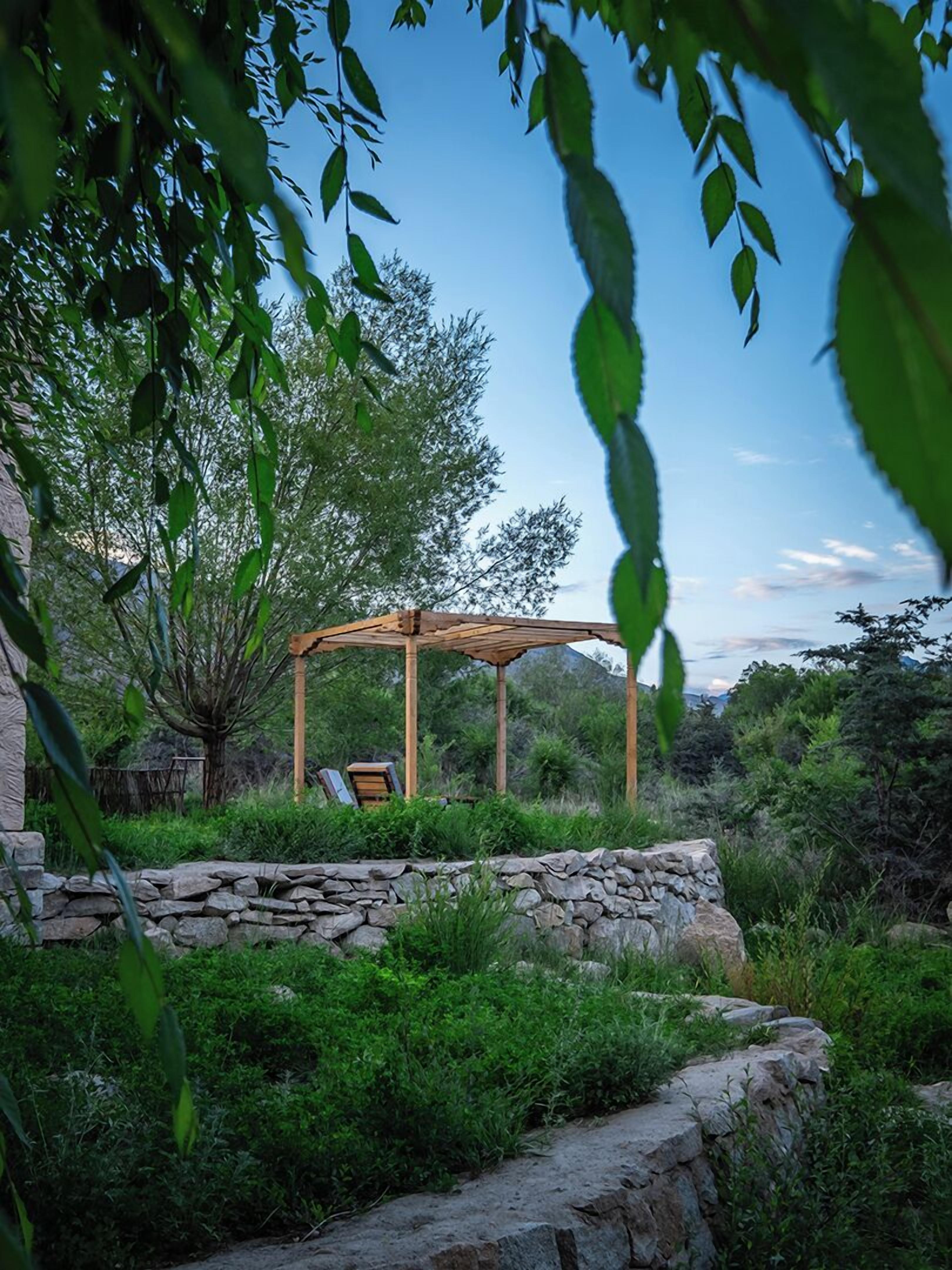 A stone wall, a wooden pergola, and a bench in a garden.