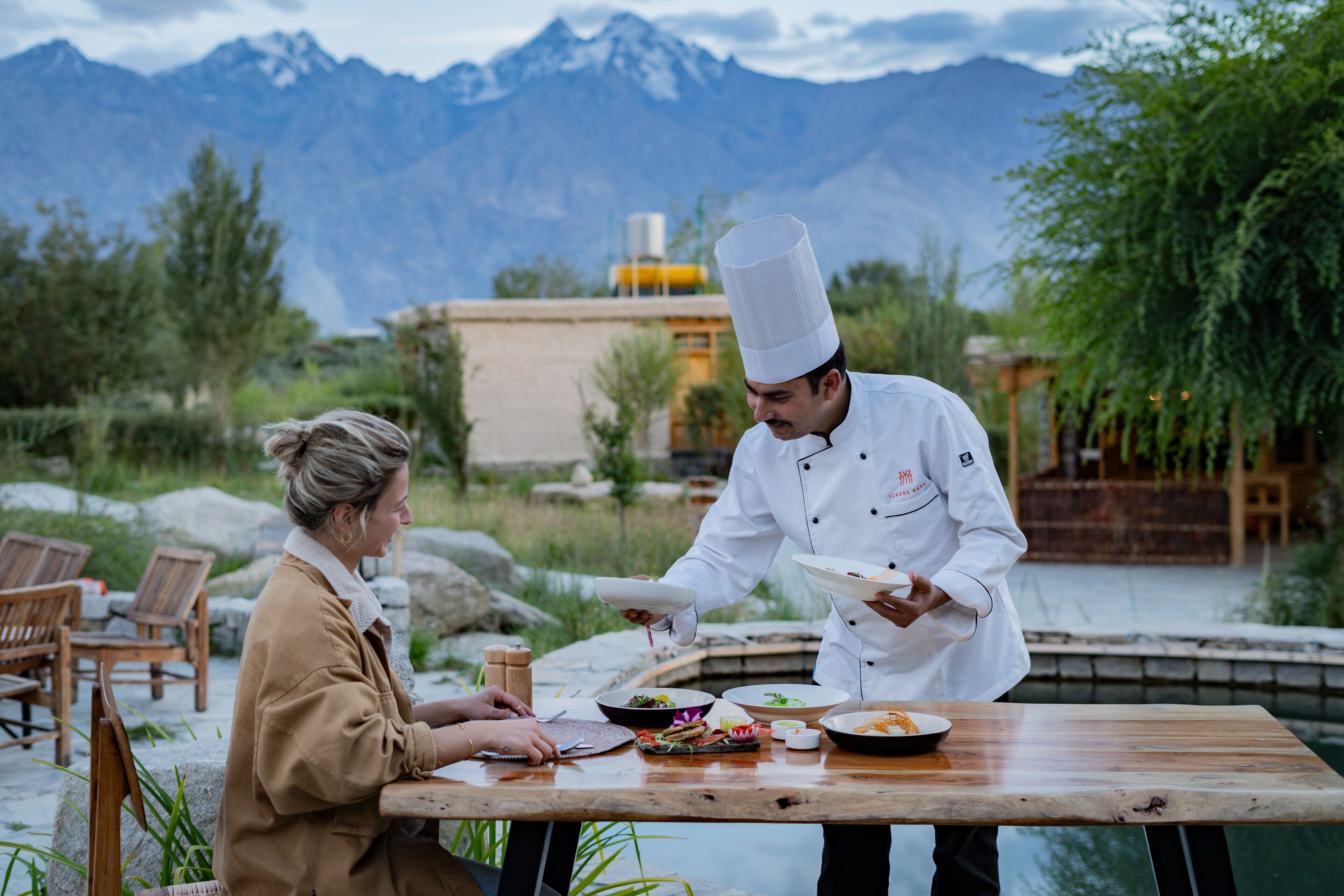 A chef serving food to a guest at an outdoor table with a mountain view in the background.