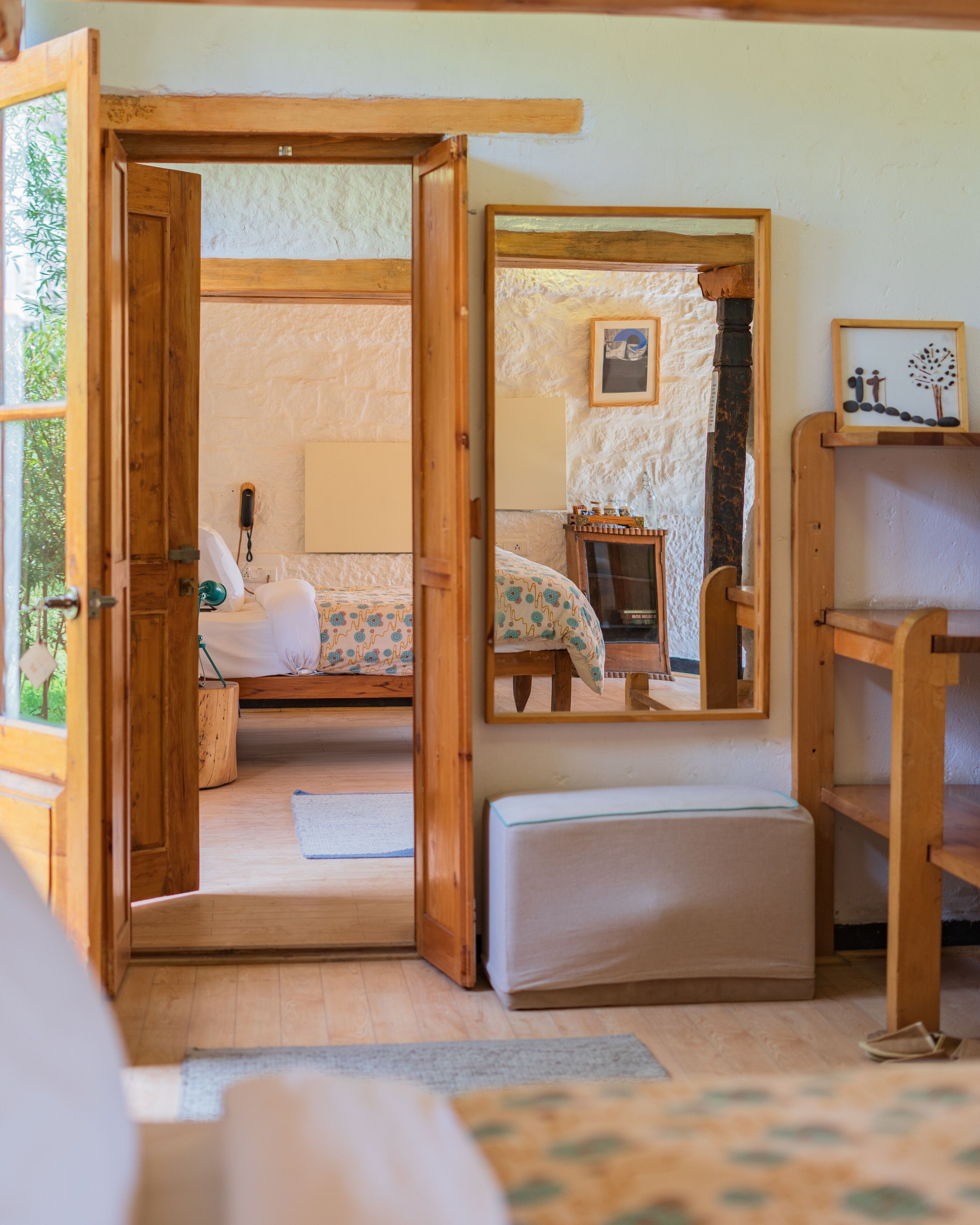 A cozy room with a wooden door leading to a bathroom, a bed with patterned bedding, a bedside table, and a mirror reflecting the room.