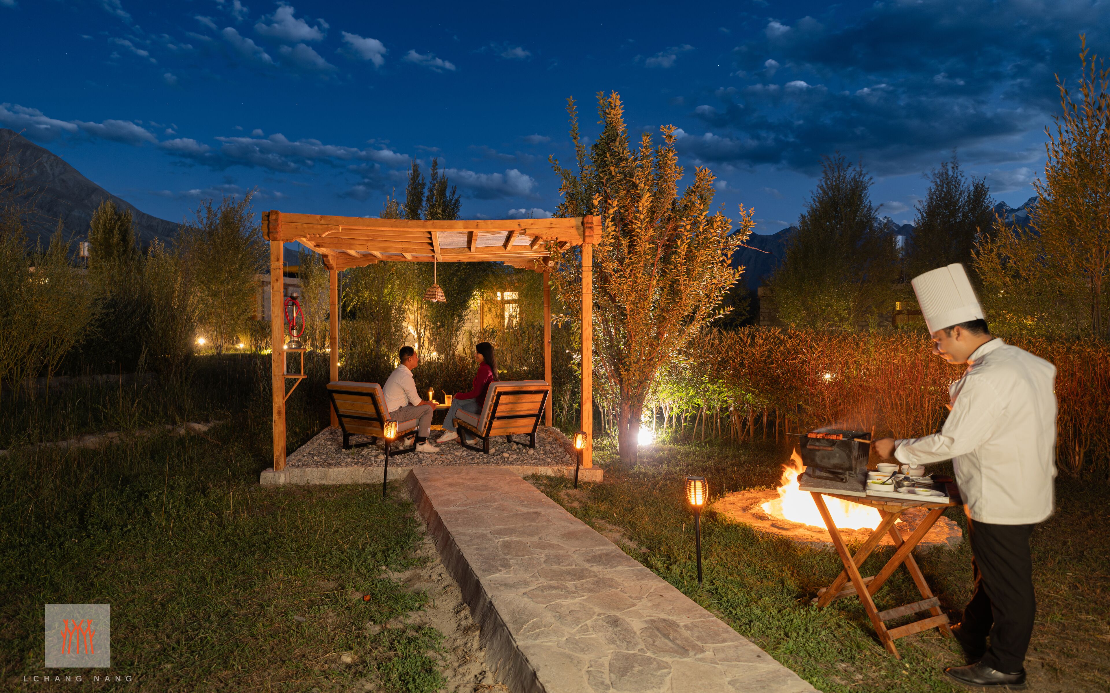 An outdoor dining area with a pergola, stone pathway, and a chef preparing food.