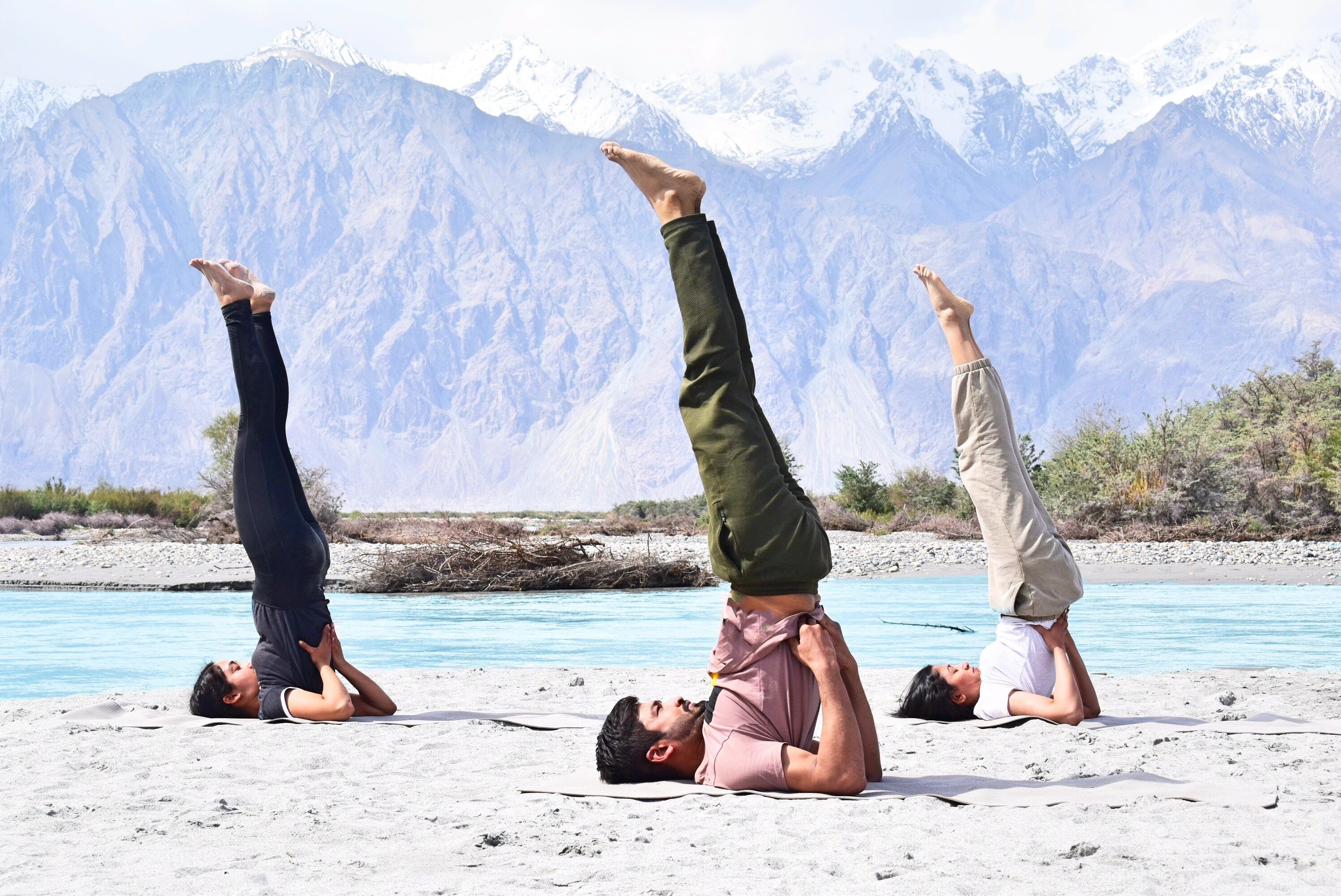 Tres personas realizando poses de yoga de parada de manos en una playa con montañas al fondo.