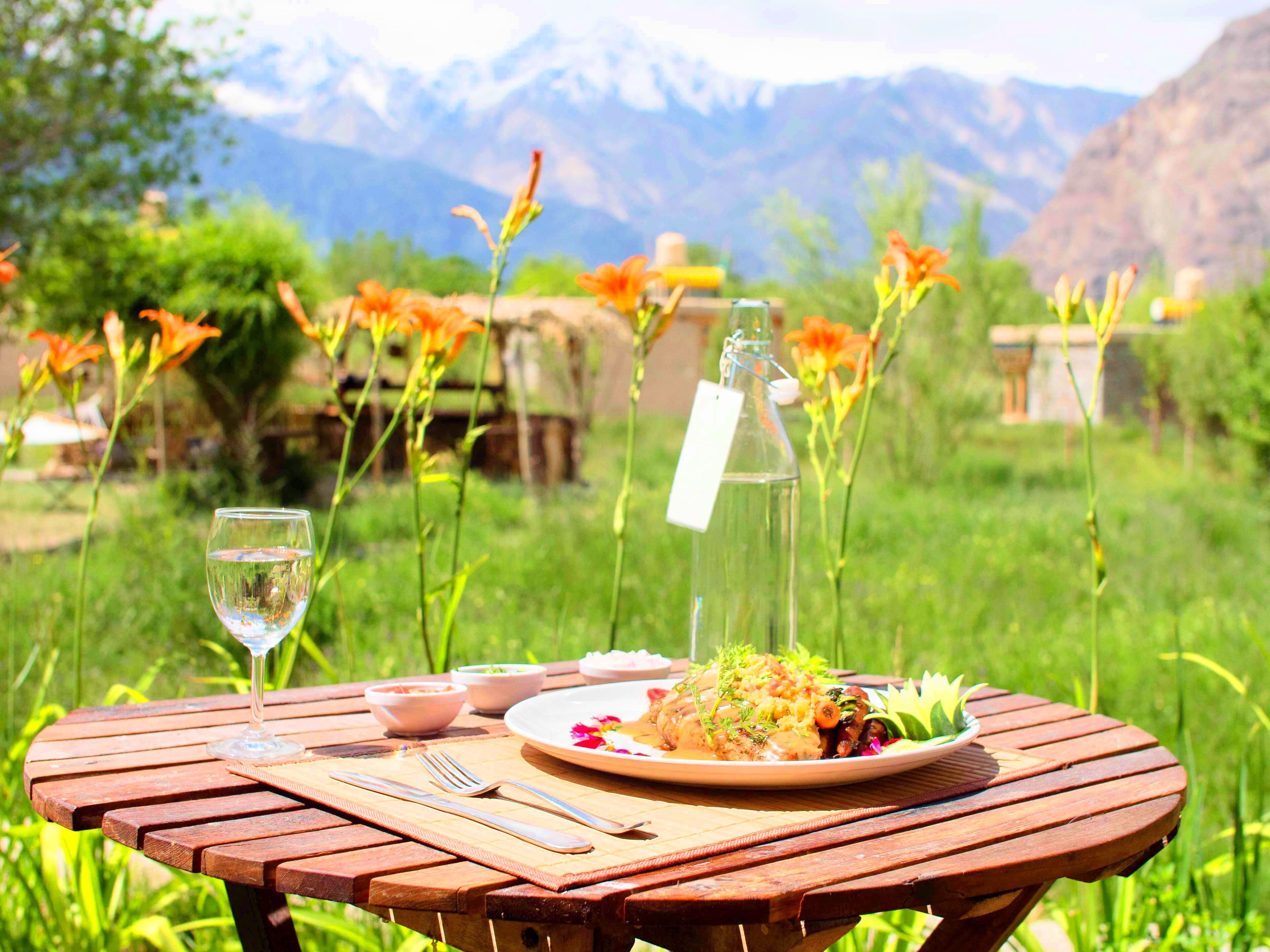 A wooden table set outdoors with a plate of food, a glass of water, and a bottle with a tag.