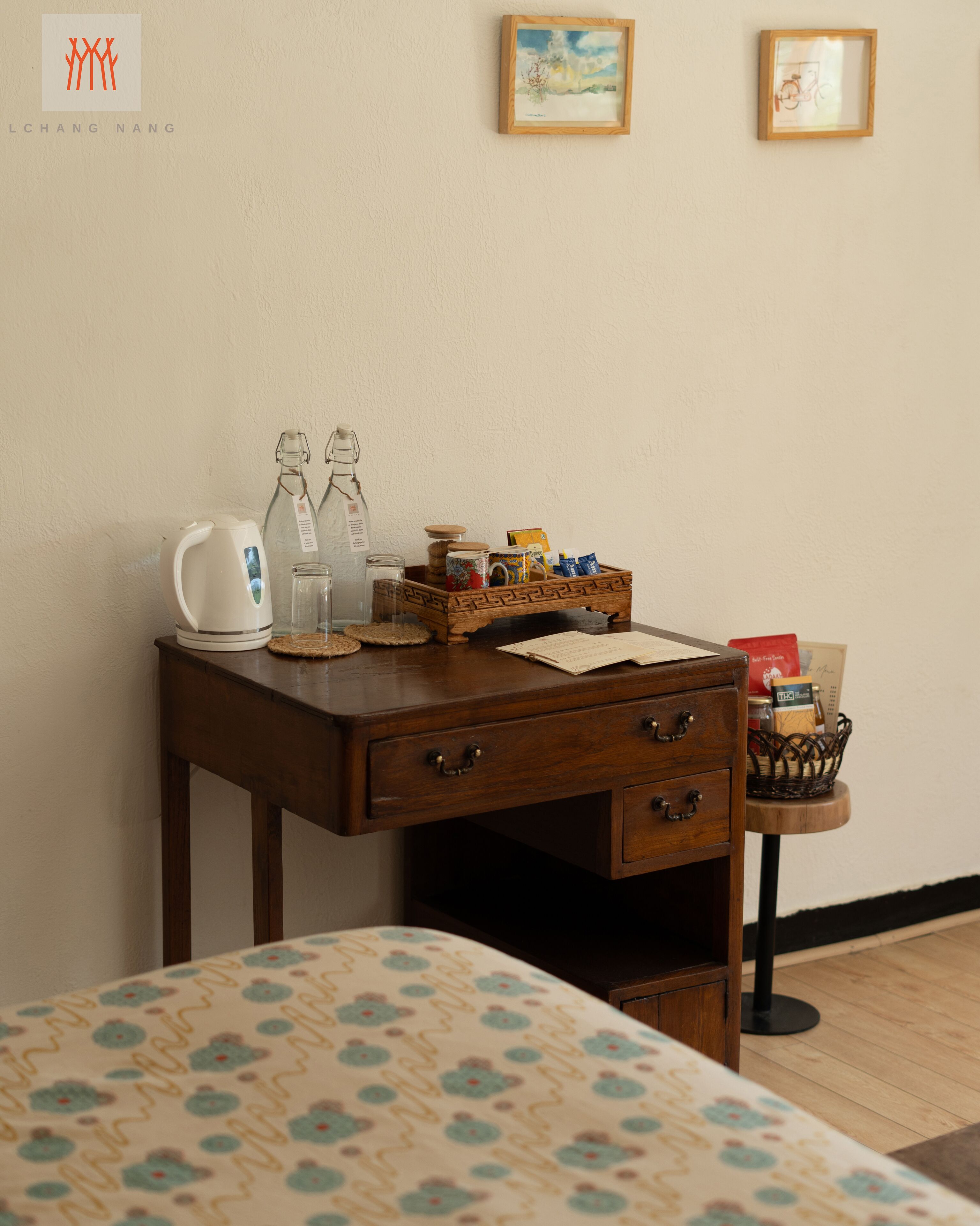Een verzorgde hotelkamer met een houten bureau, een bed met een bloemenprint, een klein tafeltje met een mand, en twee ingelijste foto’s aan de muur.