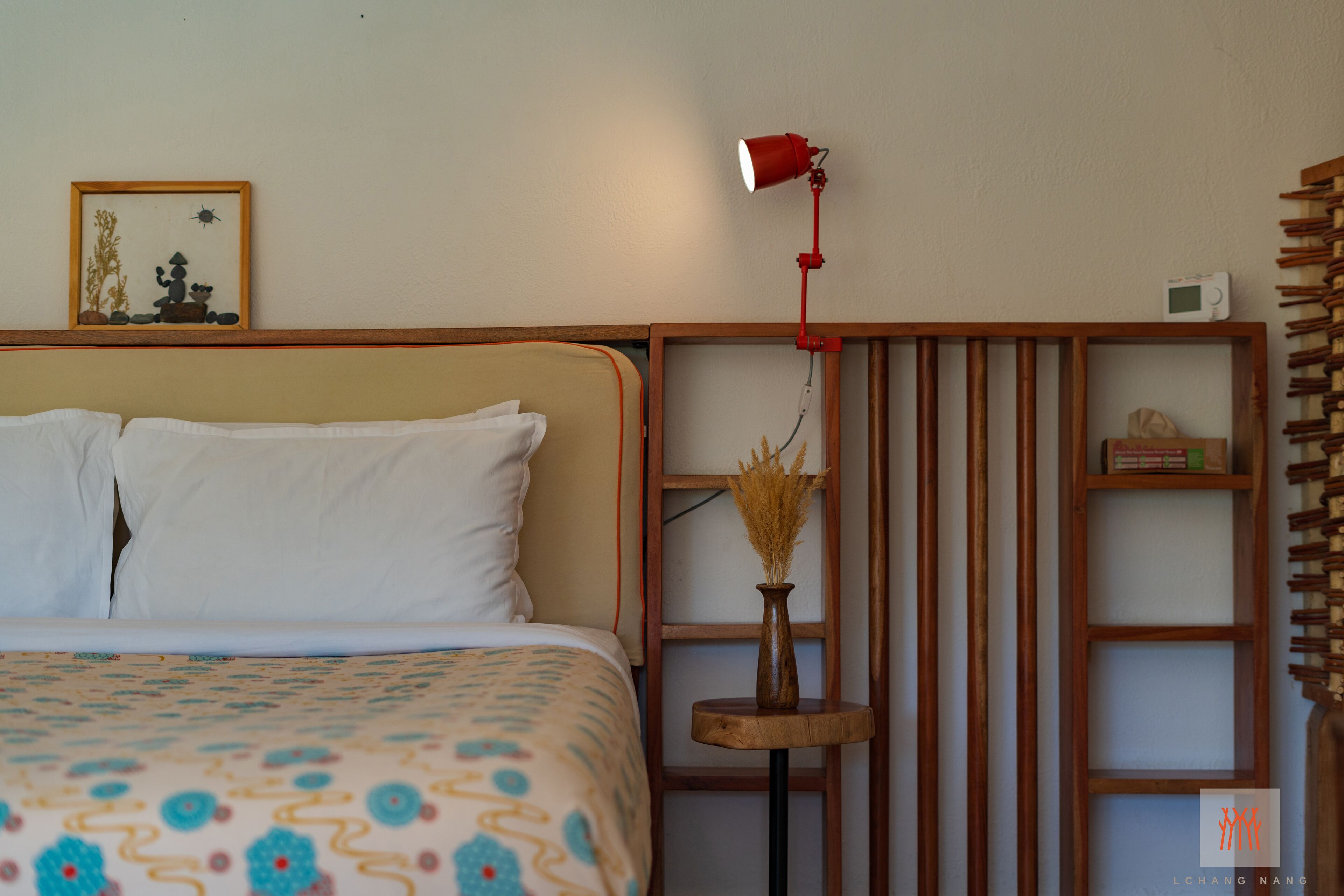A neatly made bed with a patterned bedspread, a small wooden side table with a vase, a red desk lamp, and a framed picture on the wall.
