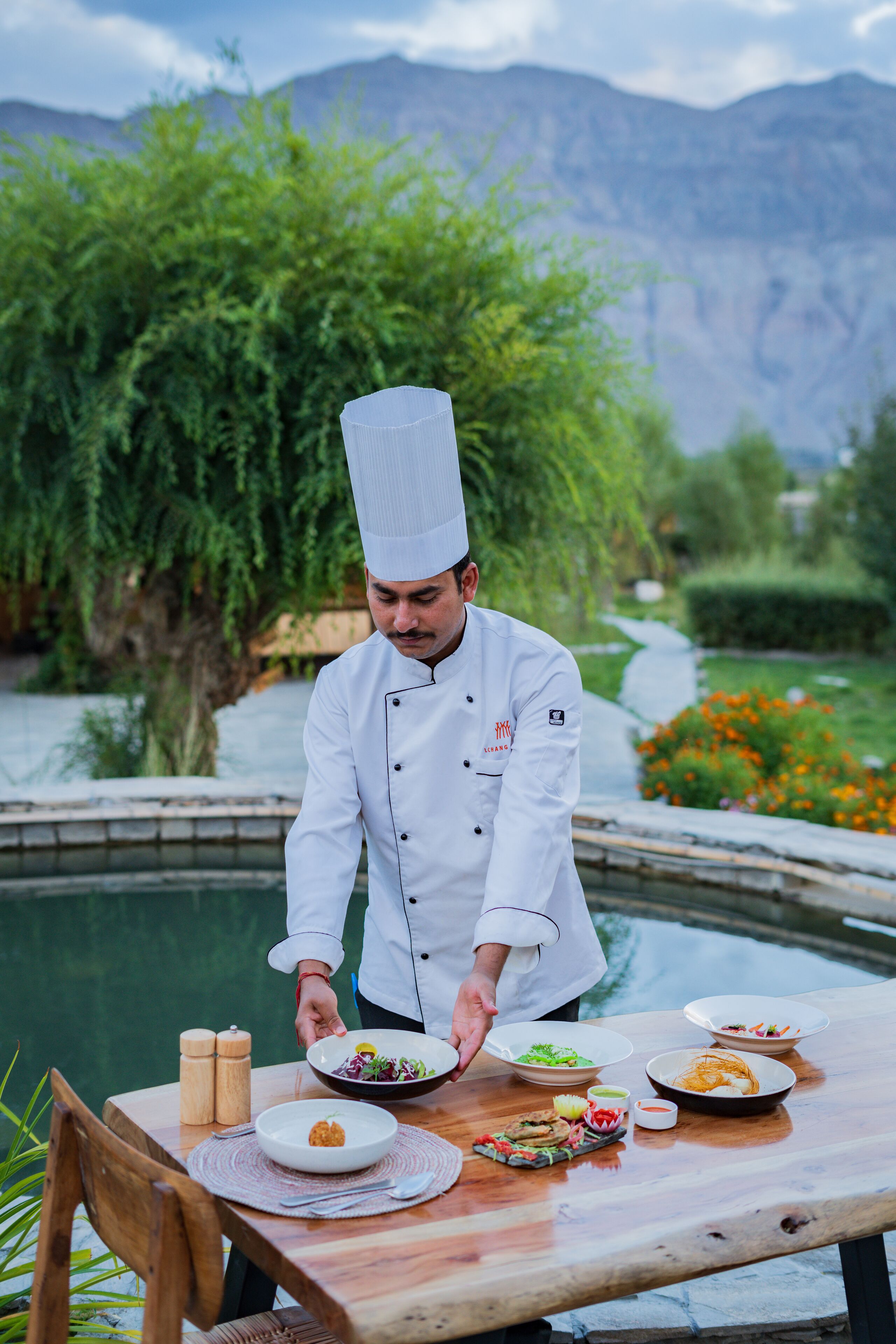A chef serving food at an outdoor table with a scenic mountain backdrop.