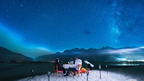 A group of people dining at a table on a beach with a starry night sky and mountains in the background.