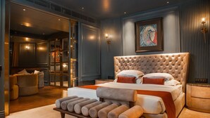 A bedroom with a tufted headboard, a large bed with multiple pillows, a bench with cushions, a framed artwork on the wall, and a sliding glass door leading to a bathroom. - Aauris (Kolkata)