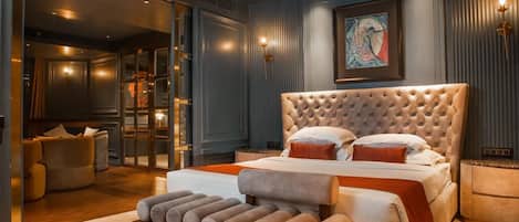 A bedroom with a tufted headboard, a large bed with multiple pillows, a bench with cushions, a framed artwork on the wall, and a sliding glass door leading to a bathroom.