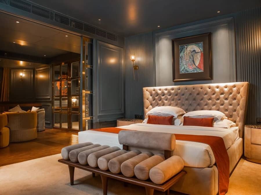 Un dormitorio con cabecera capitonada, una cama grande con muchos cojines, un banco con almohadones, una obra de arte enmarcada en la pared y una puerta corrediza de vidrio que da a un baño.