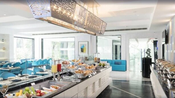 A modern hotel dining area with blue chairs, a buffet with various food items, and a large decorative light fixture. - Aauris (Kolkata)
