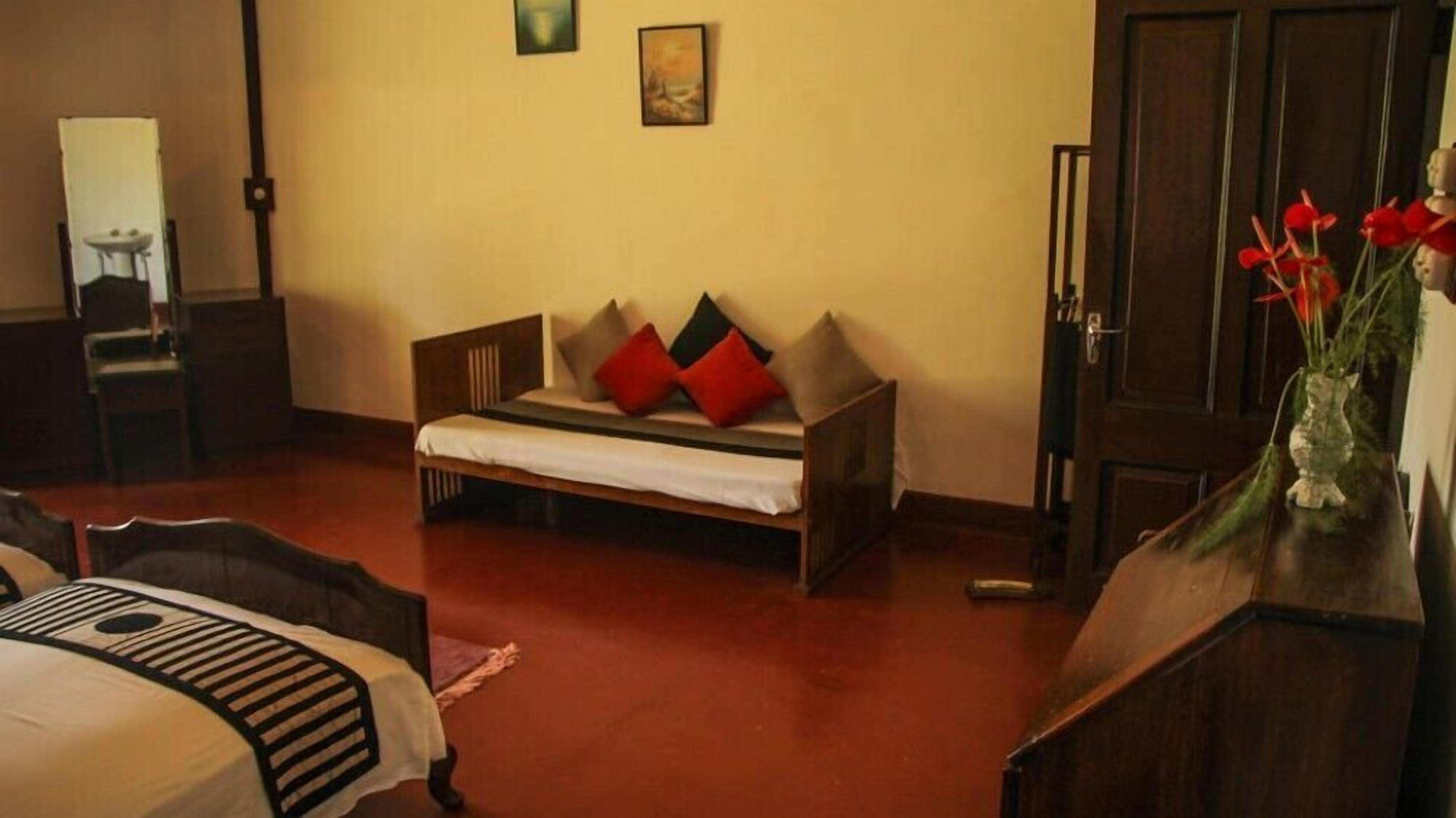Una habitación con una cama, un banco de madera, un espejo, un cuadro y un florero con flores.