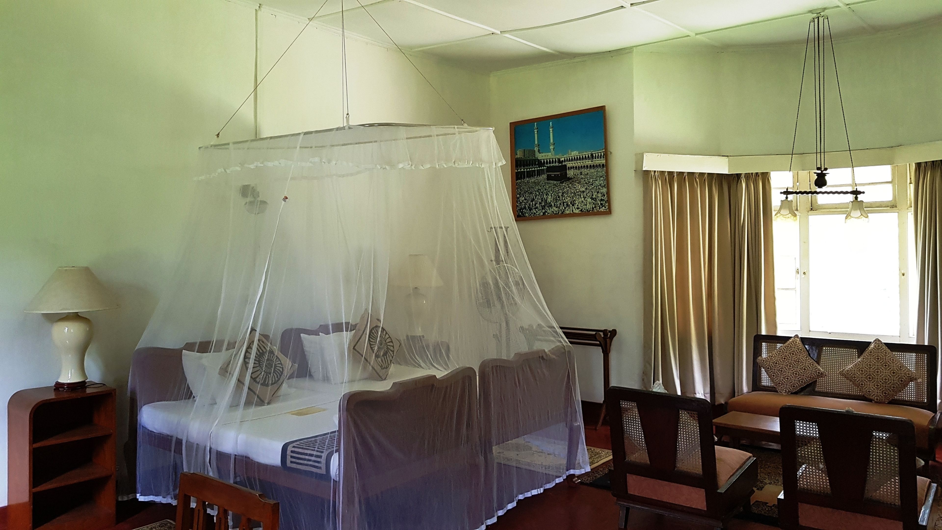 A room with a bed under a mosquito net, a sofa, a lamp, and a painting of a mosque.