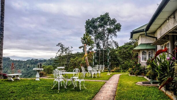Un jardin avec des chaises et des tables en métal blanc, un sentier et une maison avec un toit vert.