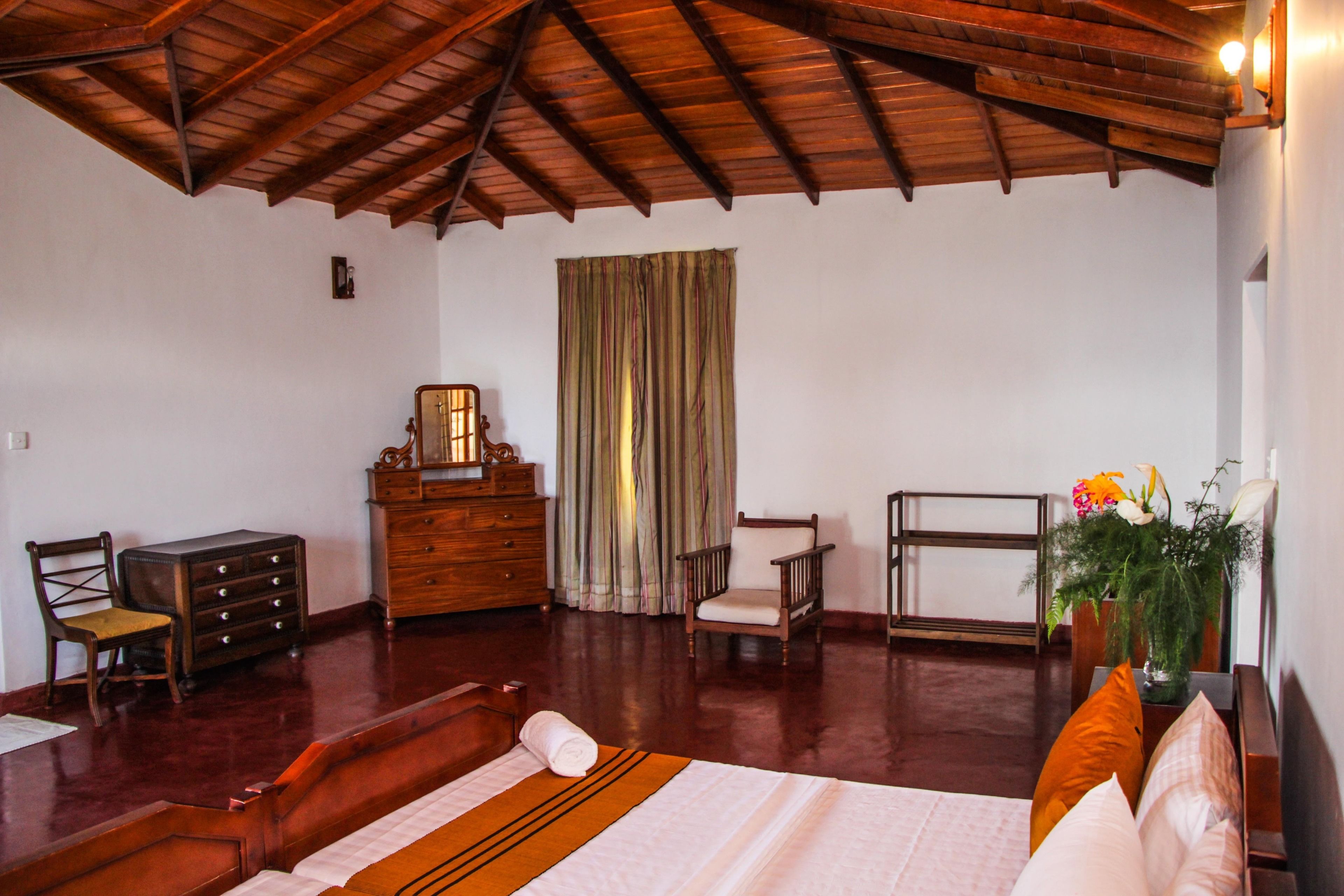 Une chambre d’hôtel avec un plafond en bois, un lit recouvert d’une courtepointe blanche, une commode en bois, une chaise, un bureau et une fenêtre avec des rideaux.