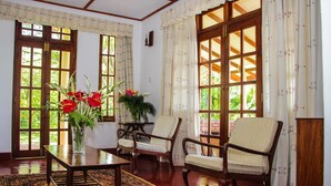 Una habitación con suelo de madera, un ventanal con vistas a zonas verdes, dos sillones tapizados, una mesa de madera y un jarrón con flores.