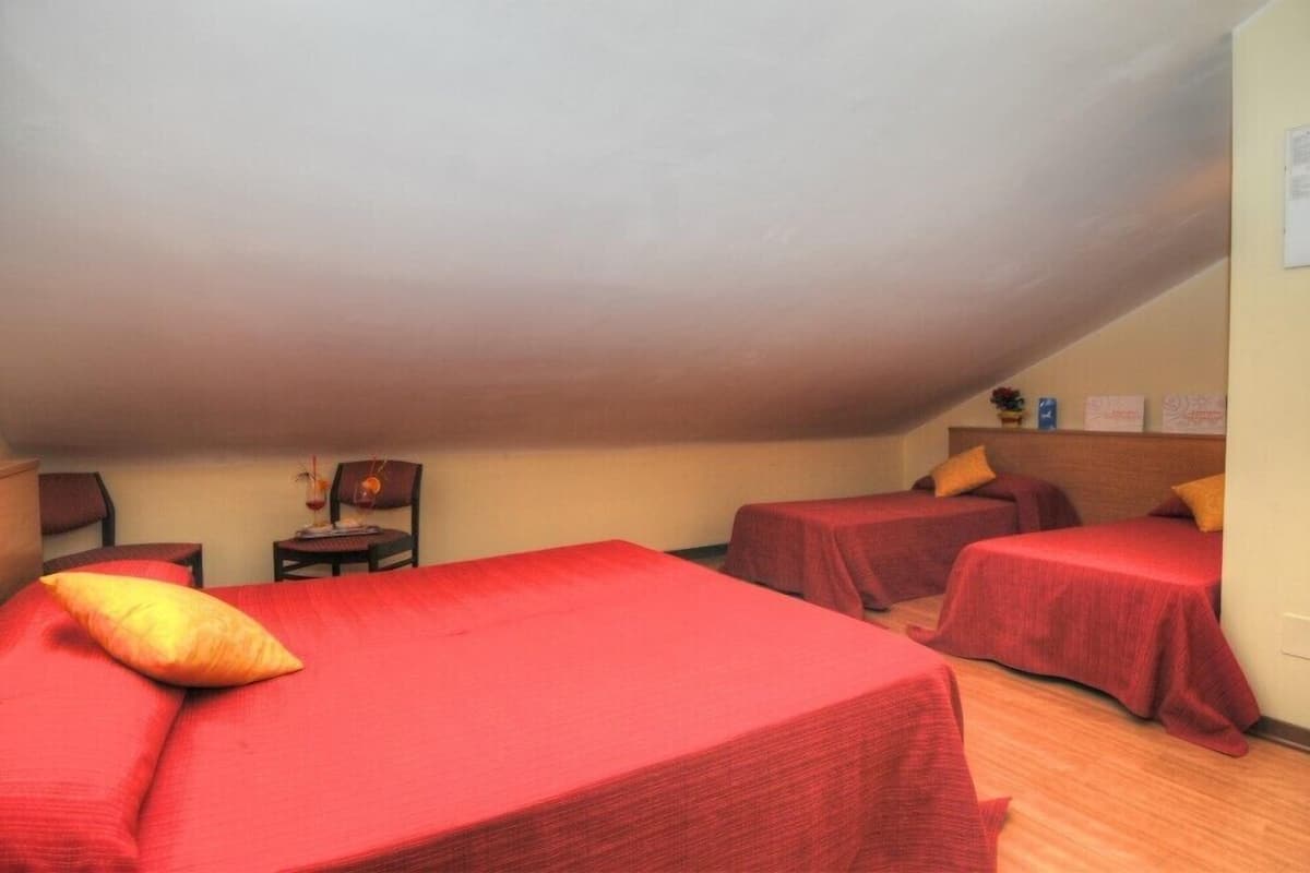 Une chambre de dortoir avec deux lits, chacun équipé de draps et d’oreillers rouges, une petite table avec une chaise et une étagère contenant divers objets.