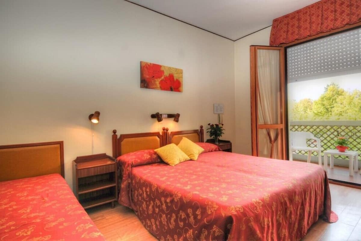 Une chambre d’hôtel avec deux lits simples, une table de chevet, une lampe, un tableau et un balcon donnant sur des arbres.