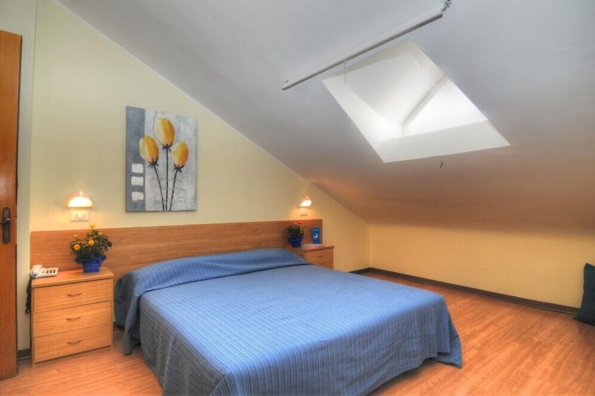 Une chambre avec une lucarne, un lit recouvert d’une courtepointe bleue, deux tables de chevet avec des lampes et un tableau accroché au mur.