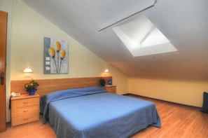 Un dormitorio con tragaluces, una cama con cubrecama azul, dos mesitas de noche con lámparas y un cuadro en la pared.