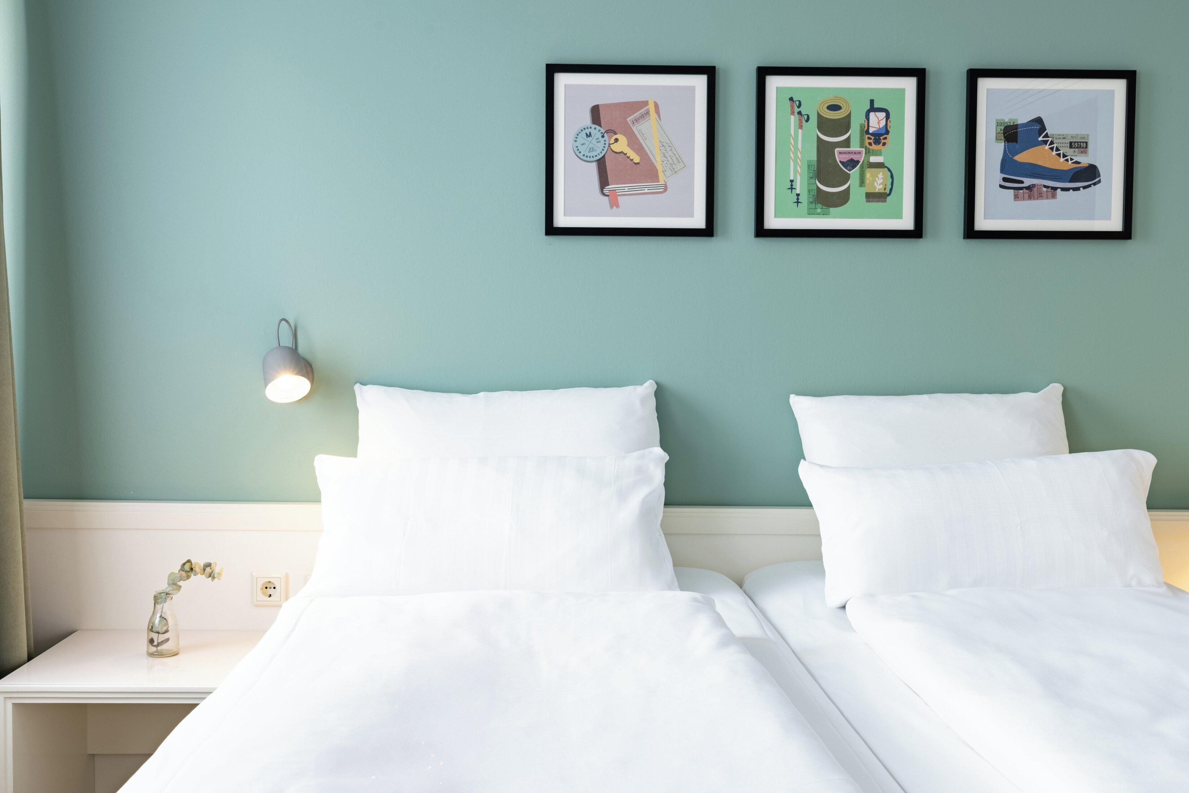 Ein Hotelzimmer mit zwei Betten, einem Nachttisch mit Lampe und drei gerahmten Bildern an der Wand.