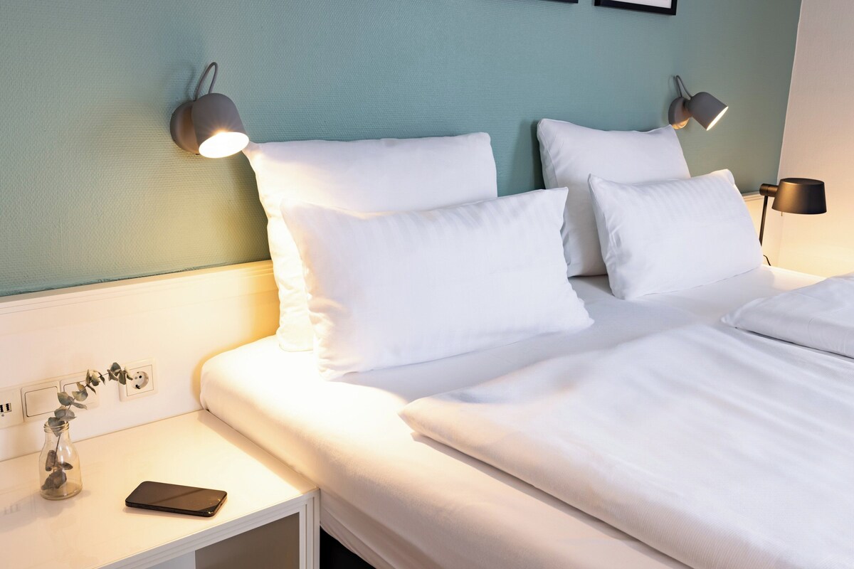 Una cama bien hecha con sábanas blancas, una mesita de noche con una lámpara y un jarrón con una planta.