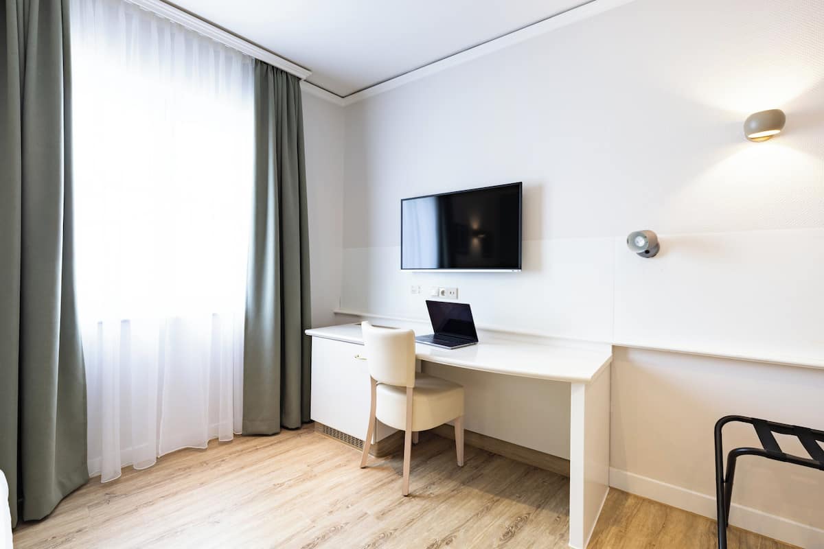 A modern room with a white desk, a laptop, a TV mounted on the wall, and a window with sheer curtains.