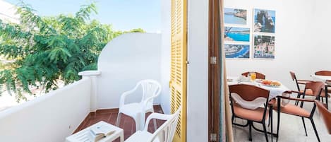 A balcony with white chairs, a table, and a dining area with a table set for two. There are framed pictures on the wall and a view of greenery and a building in the background.