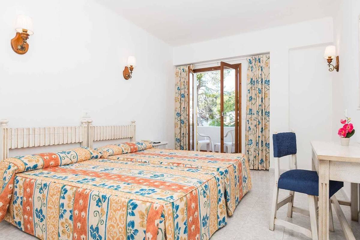 A hotel room with two beds, a desk, and a chair. There is a window with curtains and a view of the outdoors.