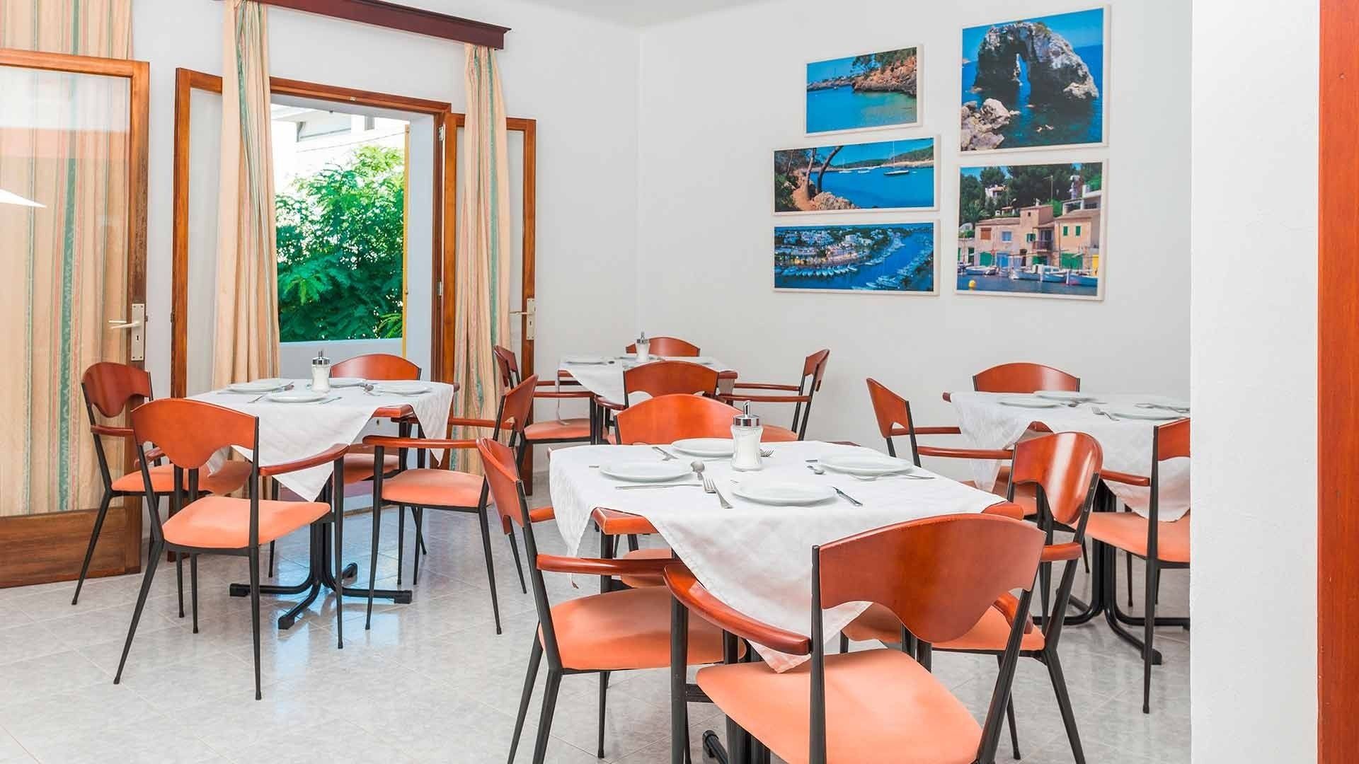 Un comedor con mesas dispuestas para cuatro, sillas anaranjadas y vistas a zonas verdes a través del ventanal.