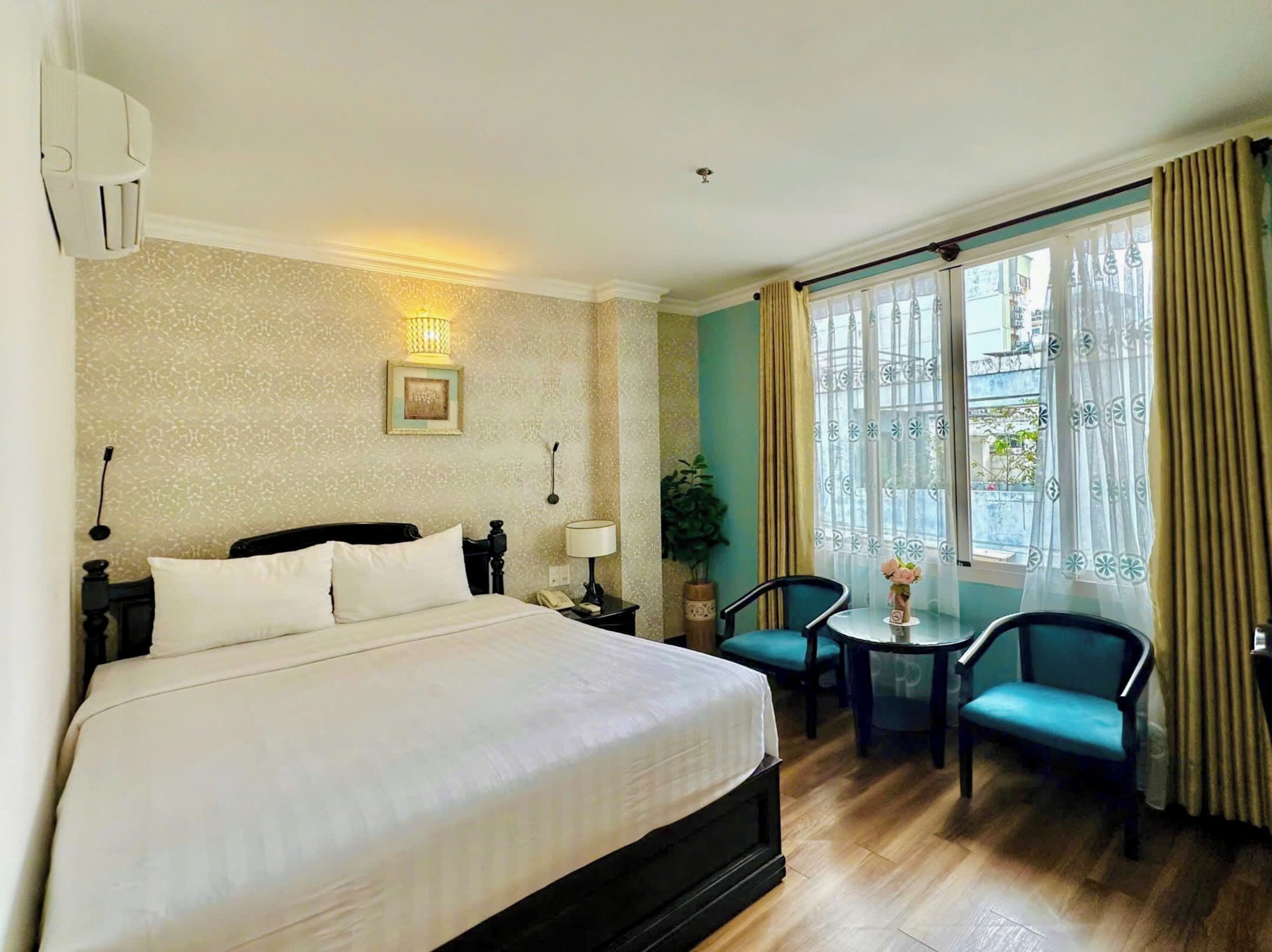 A hotel room with a large bed, a small table with chairs, a potted plant, and a window with curtains.