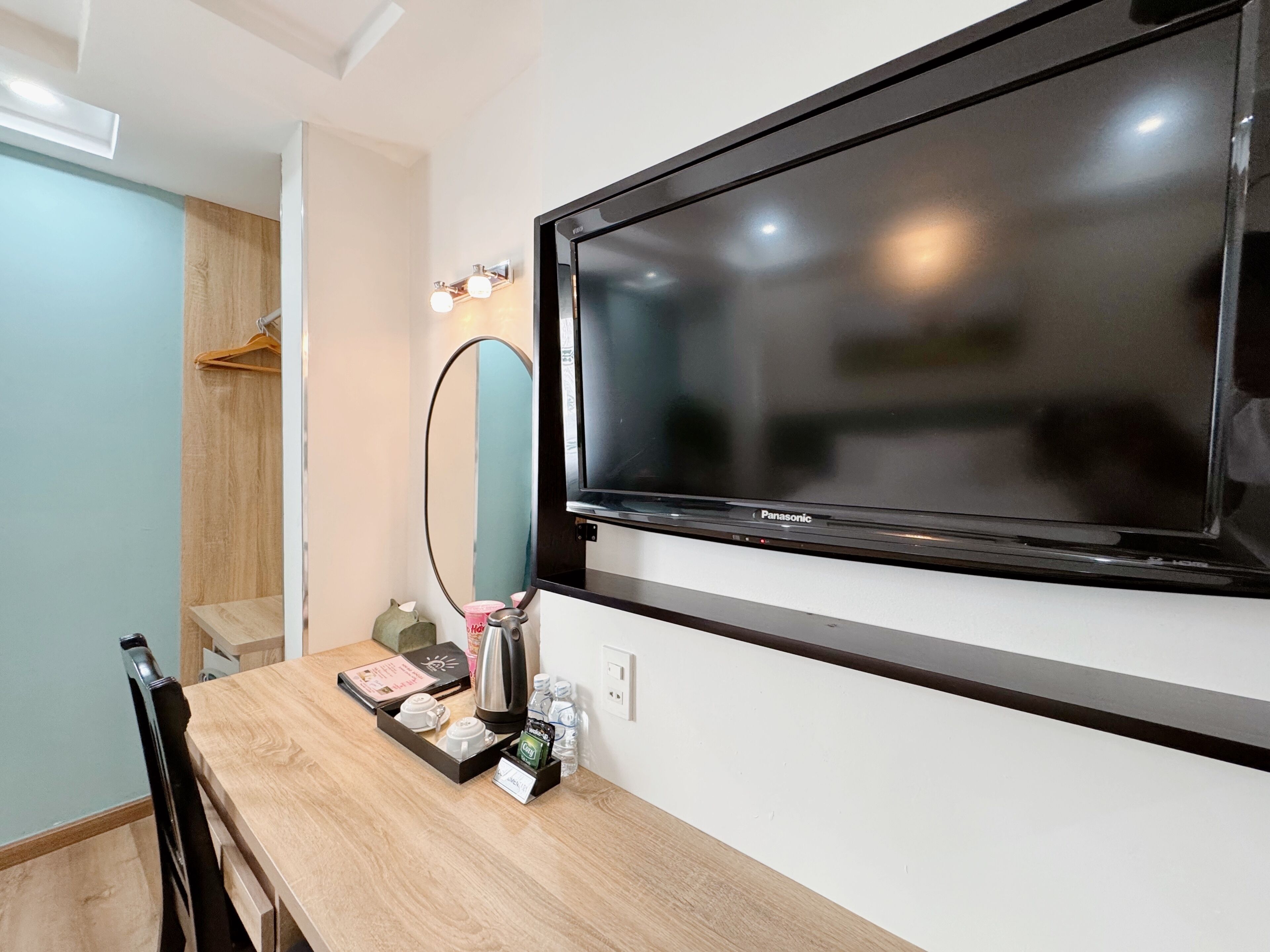 Habitación de hotel con televisor de pantalla plana montado en la pared, mesa de madera con una bandeja de alimentos para el desayuno, un espejo y un armario con ropa colgada.