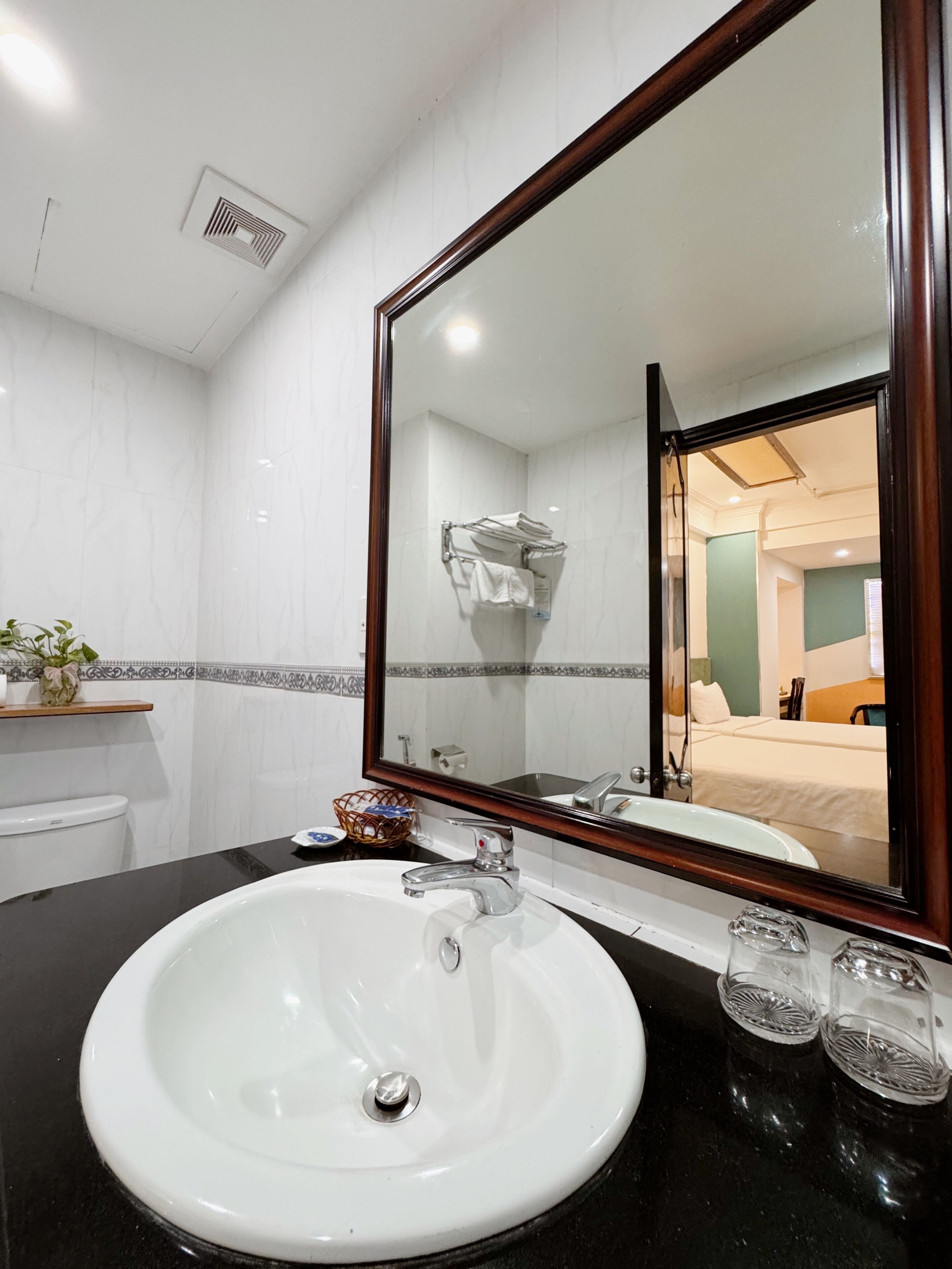 Un baño de hotel con un lavamanos blanco, un espejo grande y una vista del dormitorio en el reflejo.