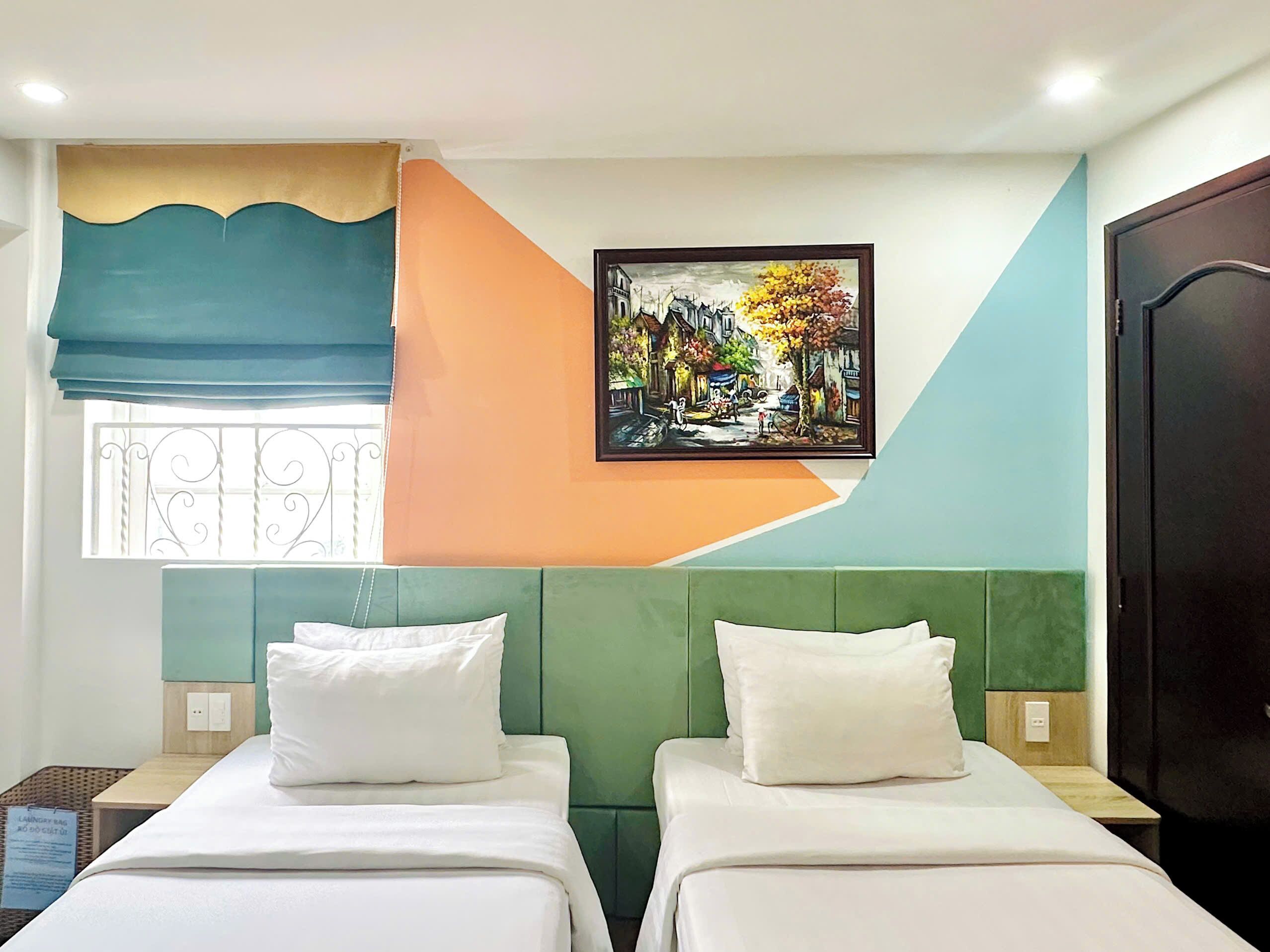 Une chambre d’hôtel avec deux lits, un tableau représentant une scène de rue, une fenêtre avec un rideau et une porte.