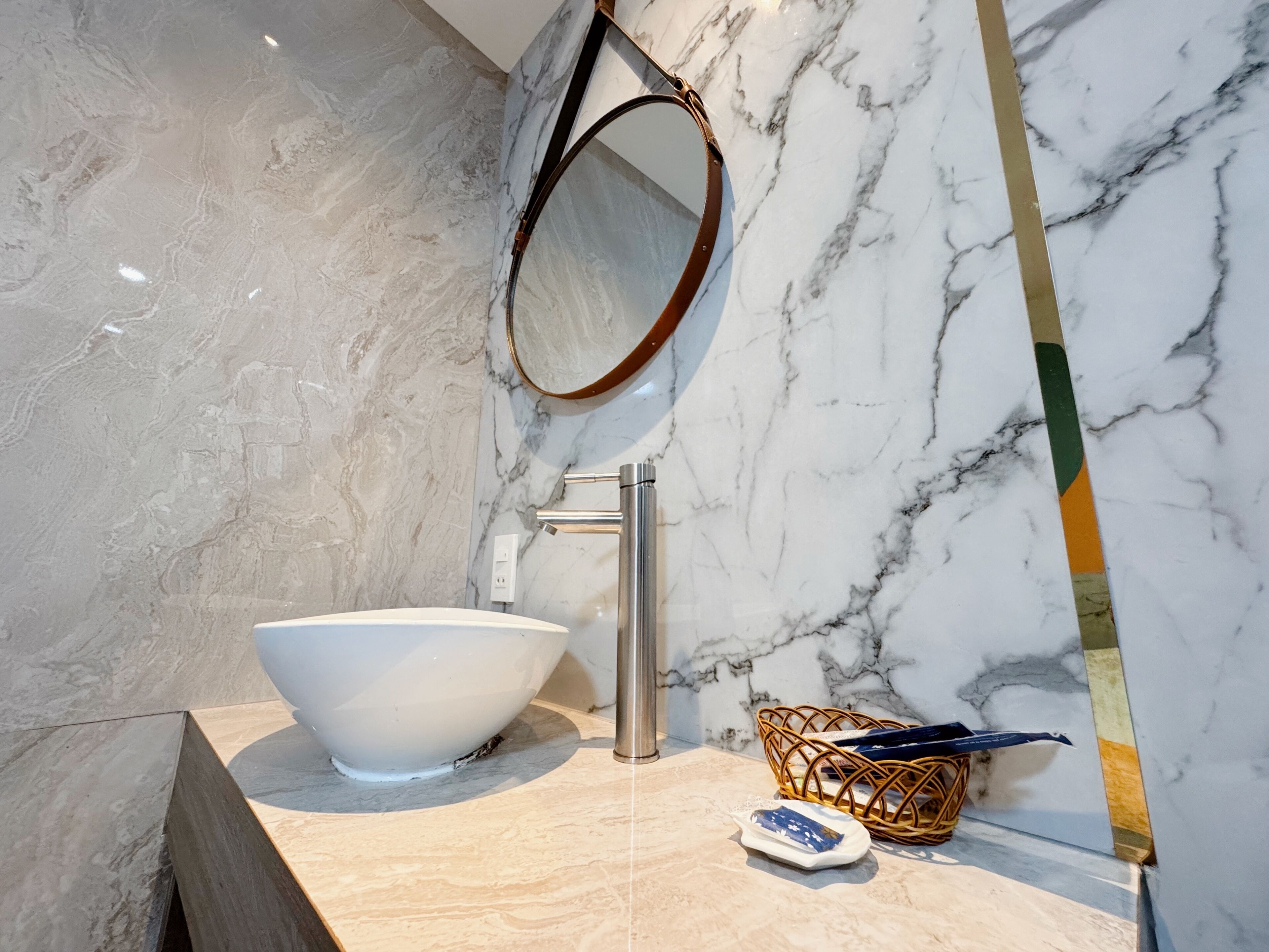 A bathroom with a white marble sink, a chrome faucet, a round mirror, and a wicker basket on the countertop.